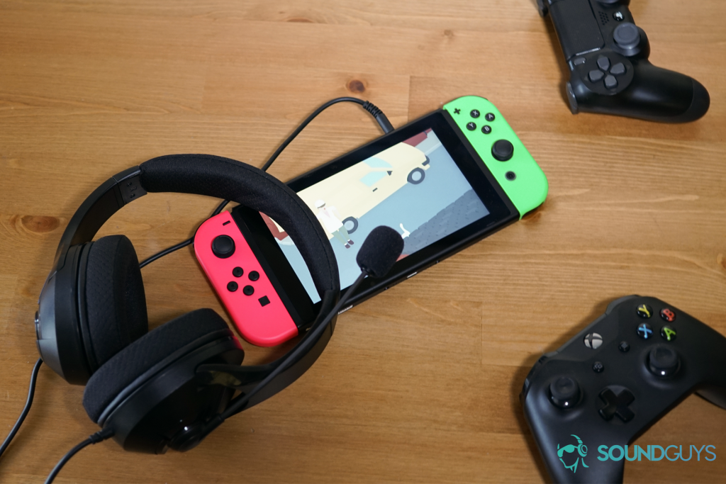 The AmazonBasics Gaming Headset leaning on a Nintendo Switch running Untitled Goose Game on a table near an Xbox One controller and Playstation 4 controller.