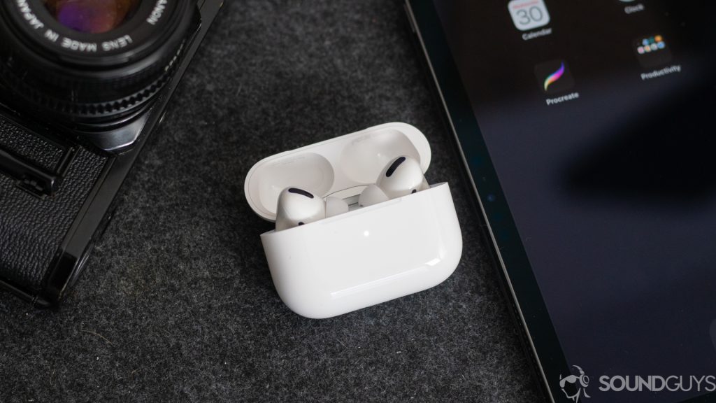 A photo of the AirPods Pro earbuds in the wireless charging case next to an iPhone and digital camera, used to illustrate build differences between the AirPods and Samsung Galaxy Buds.