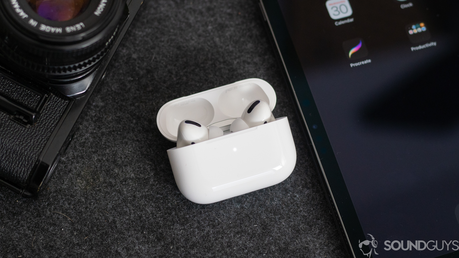 A photo of the AirPods Pro earbuds in the wireless charging case next to an iPhone and digital camera. - Apple AirPods Pro vs Sony WF-1000XM3 