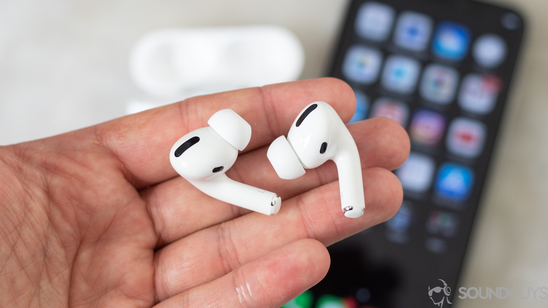 The Apple AirPods Pro in a man's left hand (foreground) with an iPhone and the AirPods Pro wireless charging case in the background.