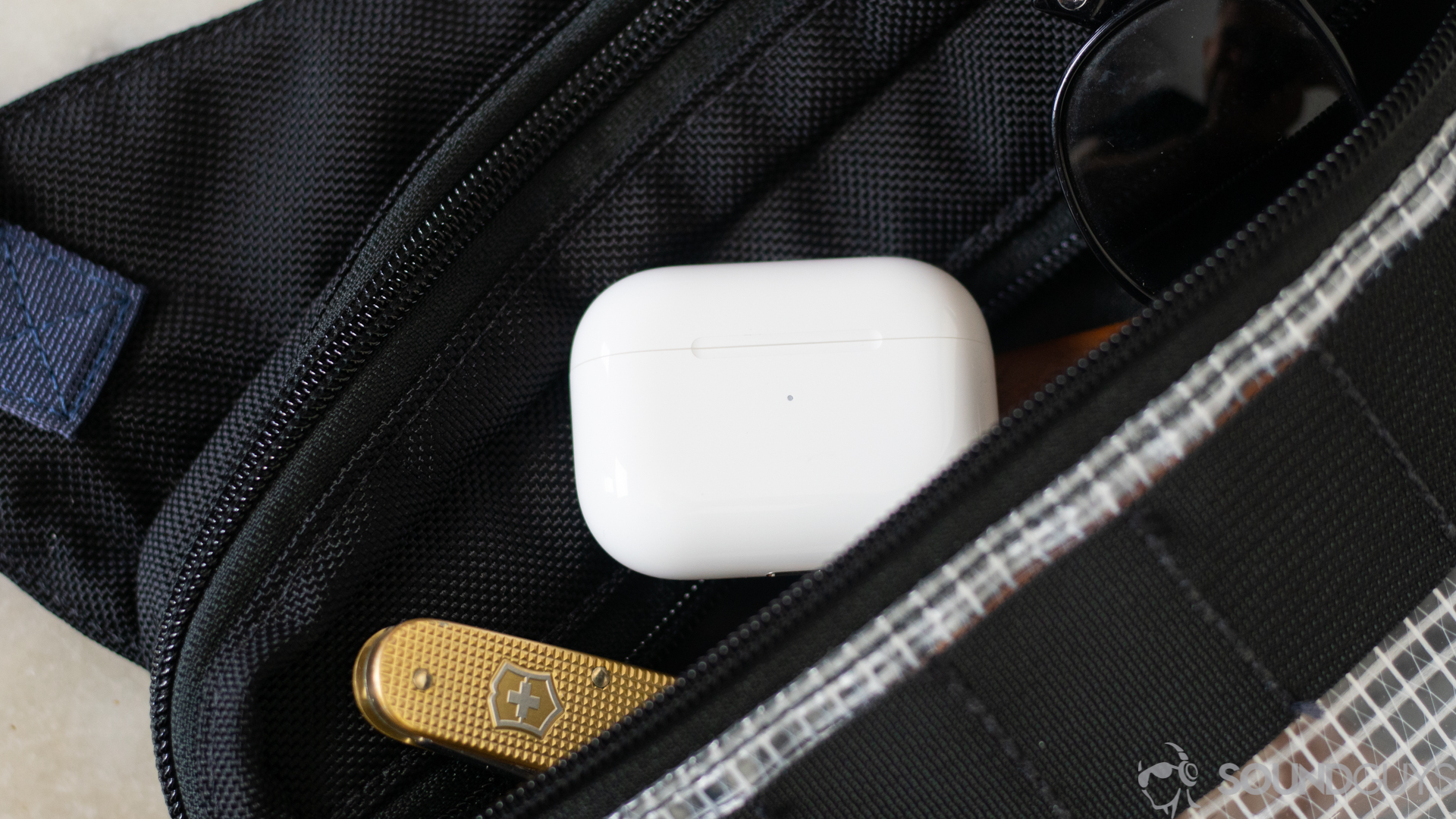 The charging case sticking out from a zippered bag pocket next to sunglasses and a swiss army knife. 