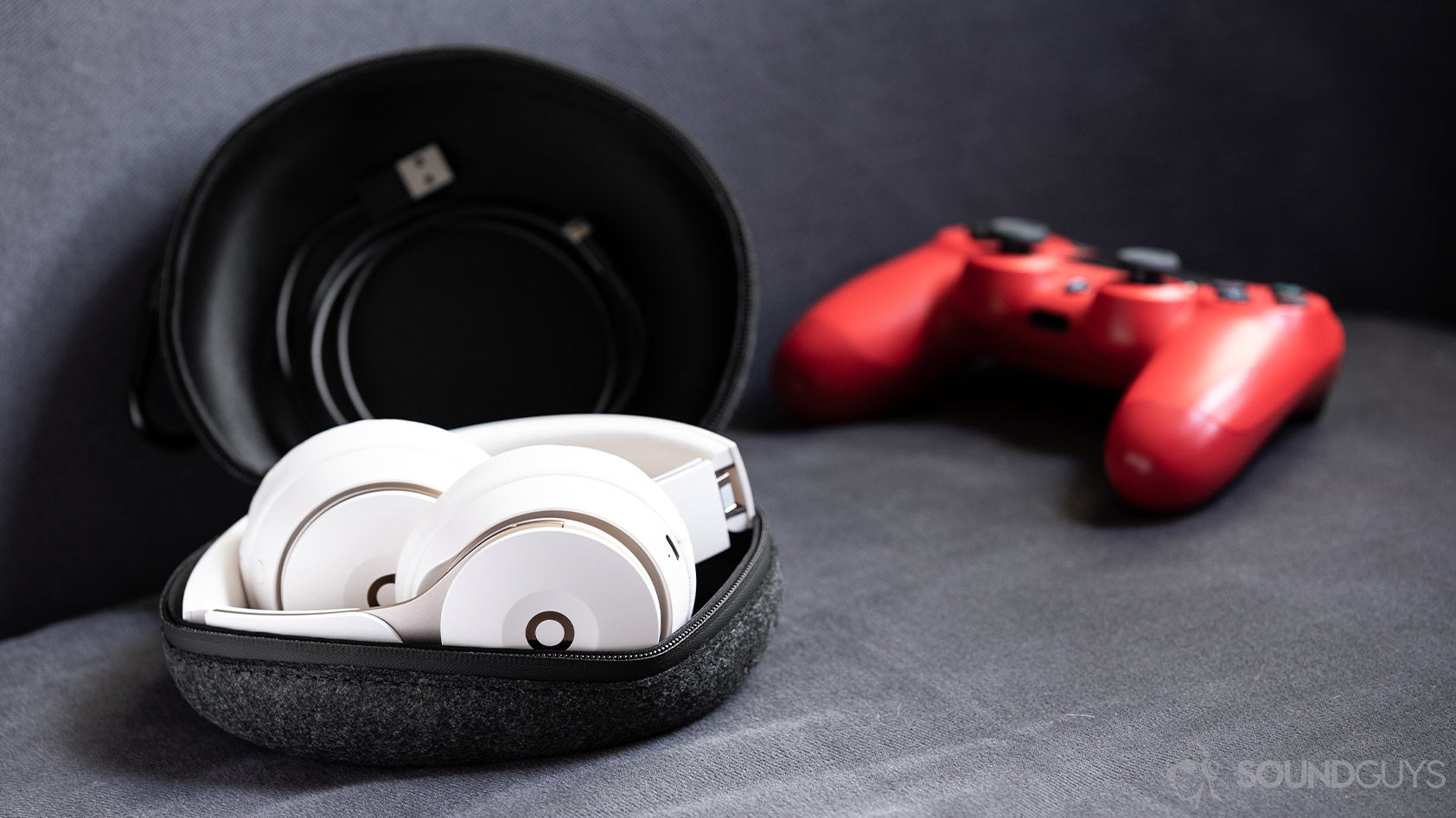 A photo of the Beats Solo Pro on-ear noise canceling headphones folded in the cloth carrying pouch with the included Lightning cable and a PS4 controller (red) in the background.