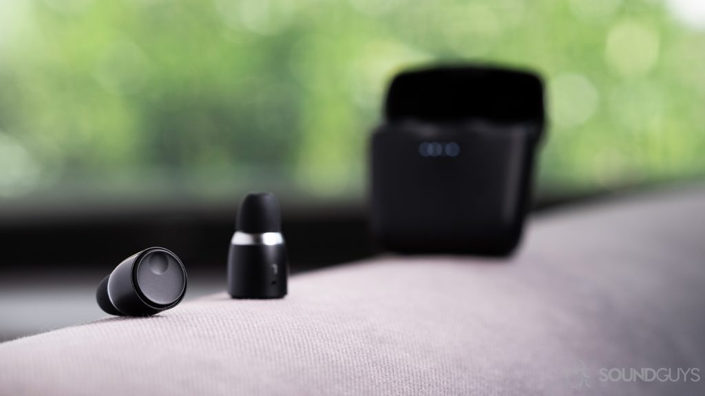 Cambridge Audio Melomania 1 true wireless earbuds outside of the case and in focus. The charging case is out of focus and open in the background. Both objects are on a cloth surface.