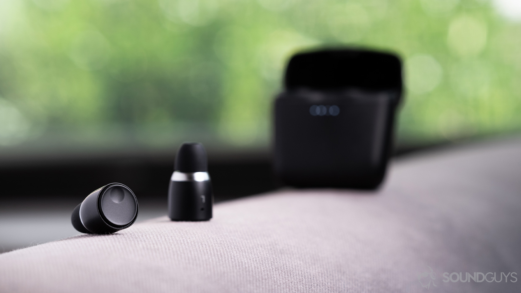 An image of the Cambridge Audio Melomania 1 true wireless earbuds outside of the case and in focus. The charging case is out of focus and open in the background. Both objects are on a cloth surface.