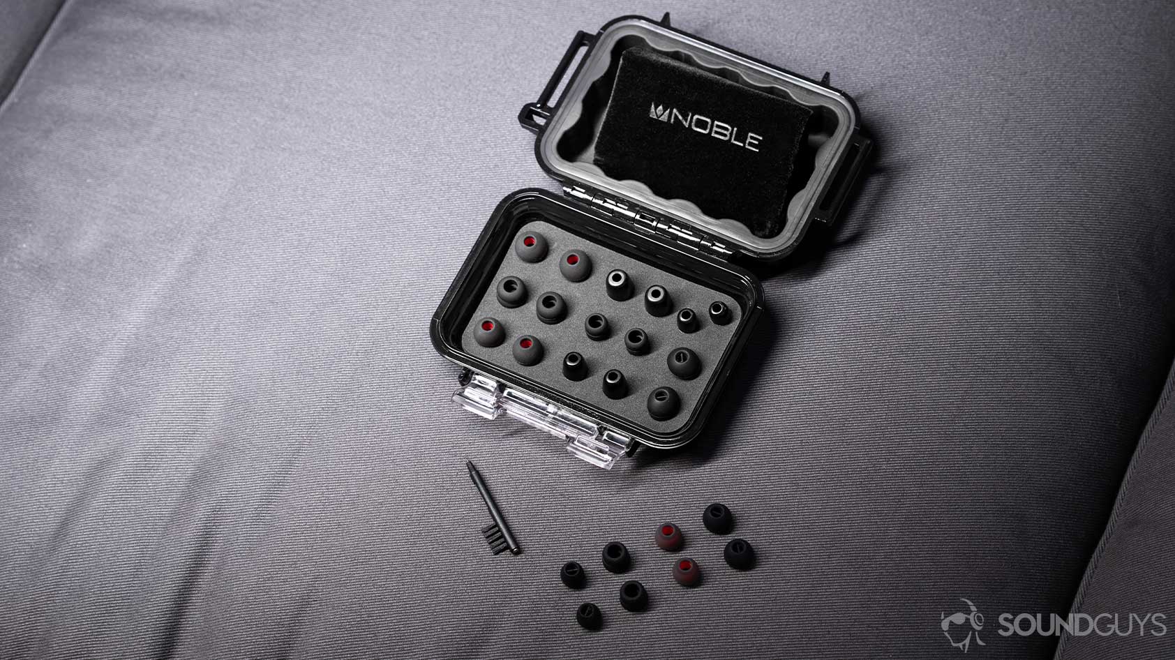 An angled, aerial photo of the Massdrop x Noble Kaiser 10 Universal Pelican carrying case open to reveal all included ear tips, an ear tip cleaner, and drawstring carrying pouch; you'd be hard-pressed to find this many accessories with any of the best true wireless earbuds under $100.
