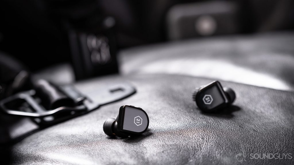 An image of the Master & Dynamic MW07 Go true wireless earbuds, which are about as large as the Samsung Galaxy Buds, on a black leather surface with an out of focus Chrome Industries messenger bag in the background.