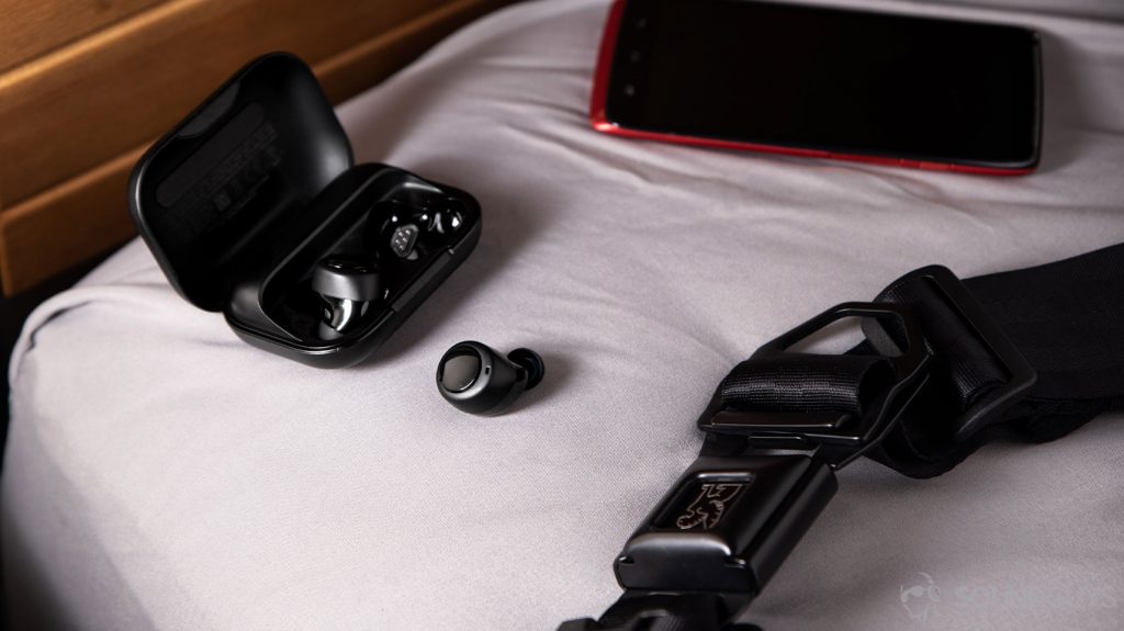 A photo of the Amazon Echo Buds with one earbud in the charging case and the other just outside of it near a Droid phone and backpack strap.