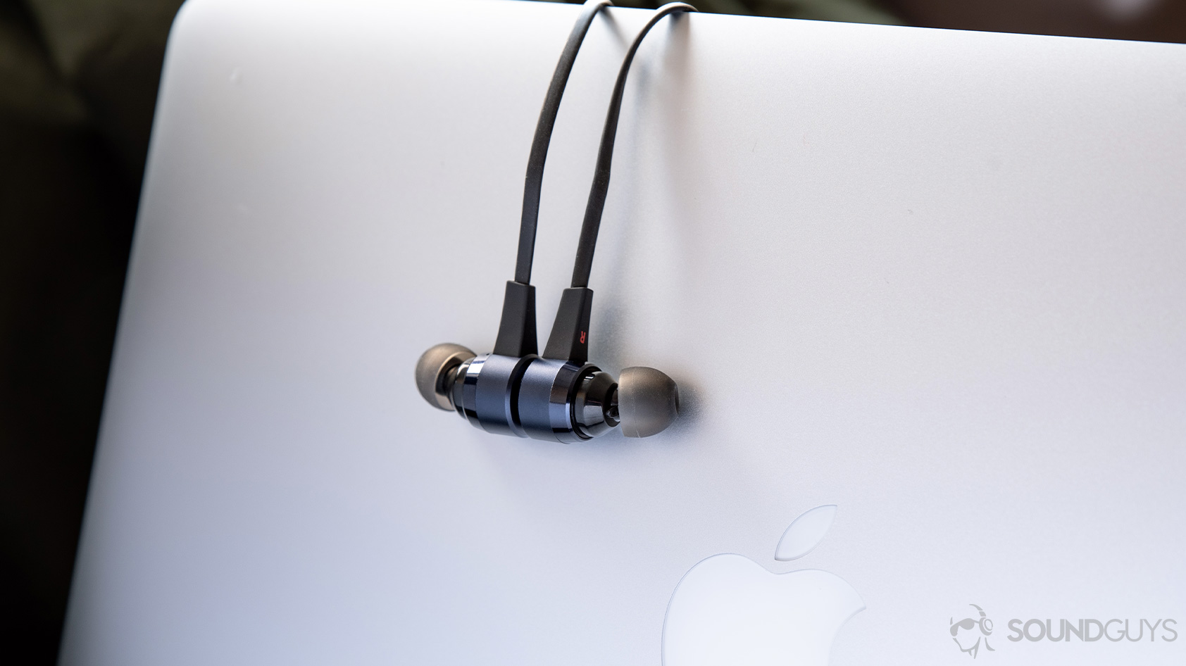 A photo of the Massdrop x NuForce Stride wireless earbuds (blue) hanging over a Macbook Pro laptop with the magnetic housings clasped together.