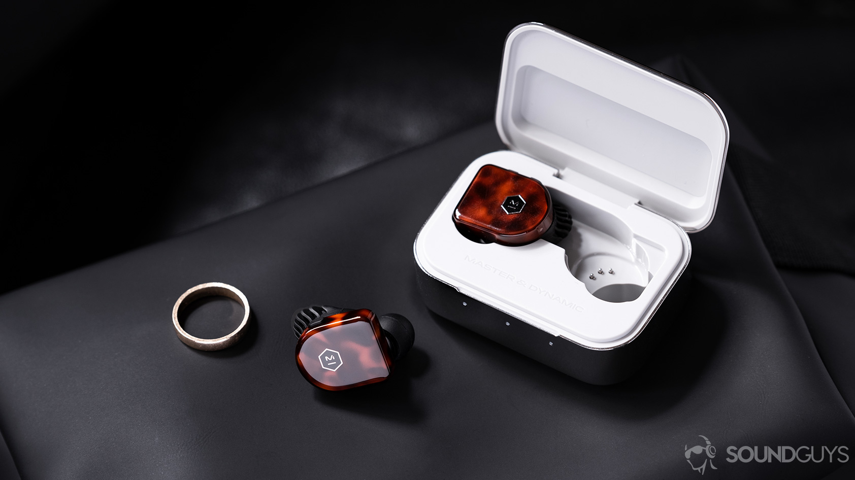 A picture of the Master &amp; Dynamic MW07 Plus noise canceling true wireless earbuds with on earbud in the case and the other outside of it on a leather surface next to a gold ring.