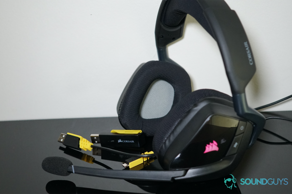 The Corsair Void RGB Elite Wireless headset sits with the cords it comes with on a black surface near a white wall.