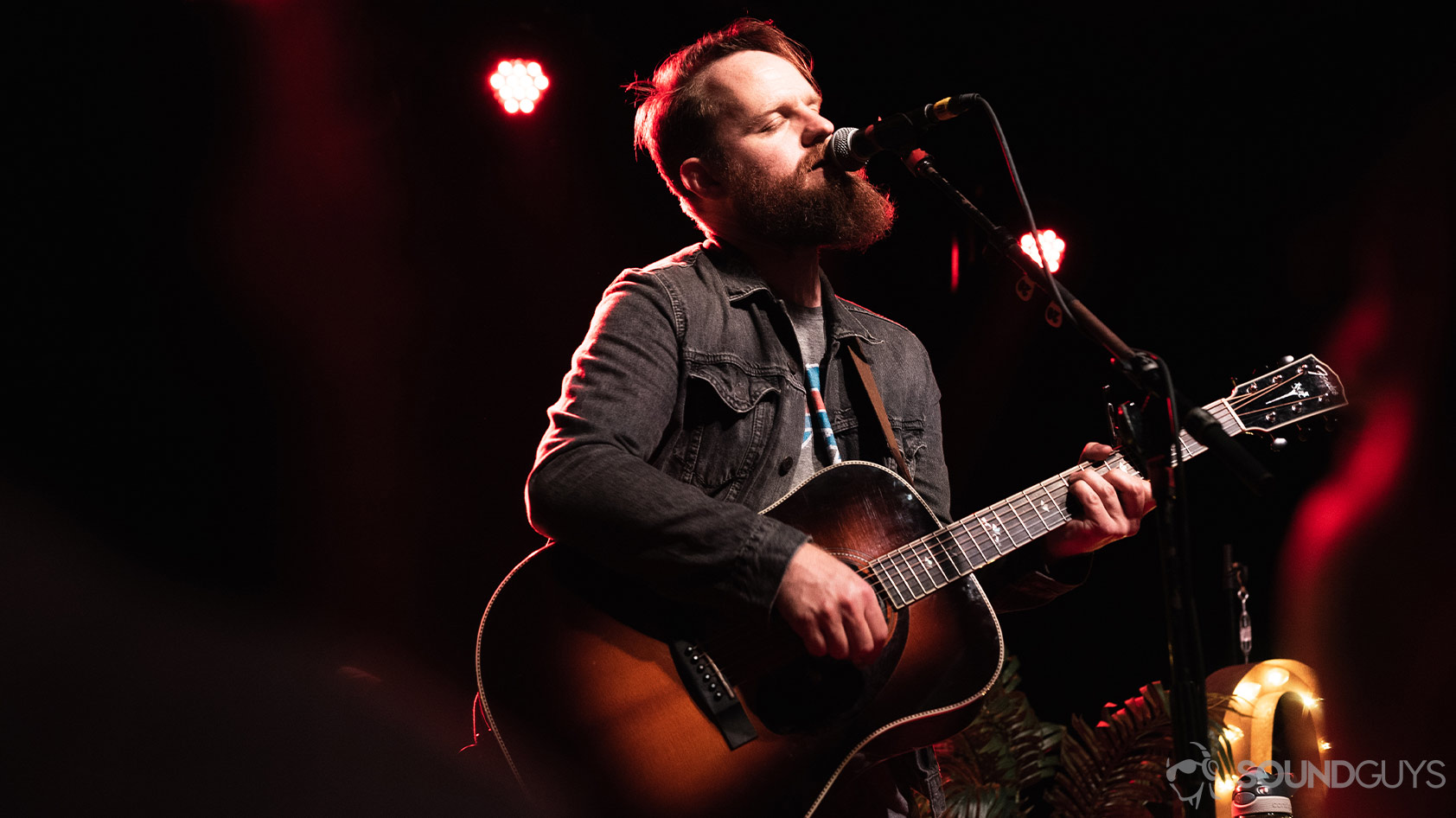 A picture of Aaron West performing with an acoustic guitar as red light is cast down on stage.