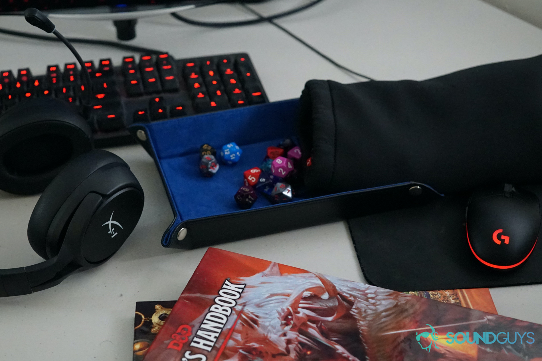 The HyperX Cloud Flight S sits on a desk in front of a computer monitor and keyboard, next to some polyhedral dice, with copies of the Dungeons and Dragons Players Handbook and Xanathar's Guide to Everything in the foreground.