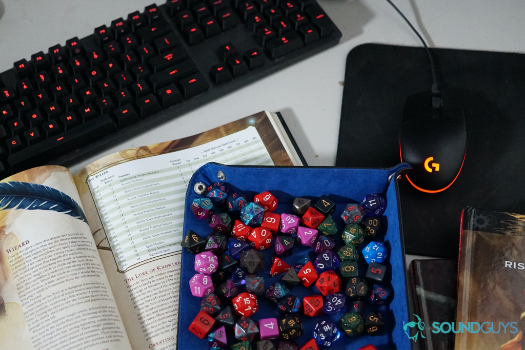 A pile of polyhedral dice sit on an open page of the Dungeons and Dragons Players Handbook in front of a Logitech mouse and keyboard, next to a copy of the Dungeons and Dragons book, Eberron: Rising from The Last War.