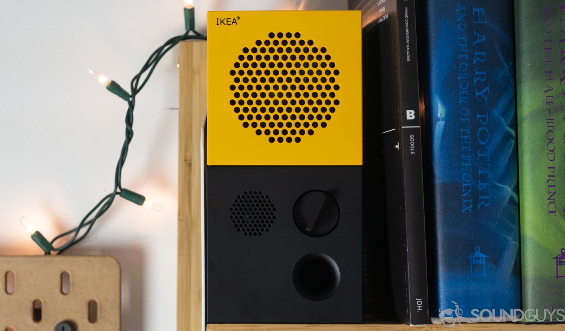The IKEA x Teenage Engineering Frekvens speaker in yellow on a bookshelf next to Harry Potter books and Christmas lights