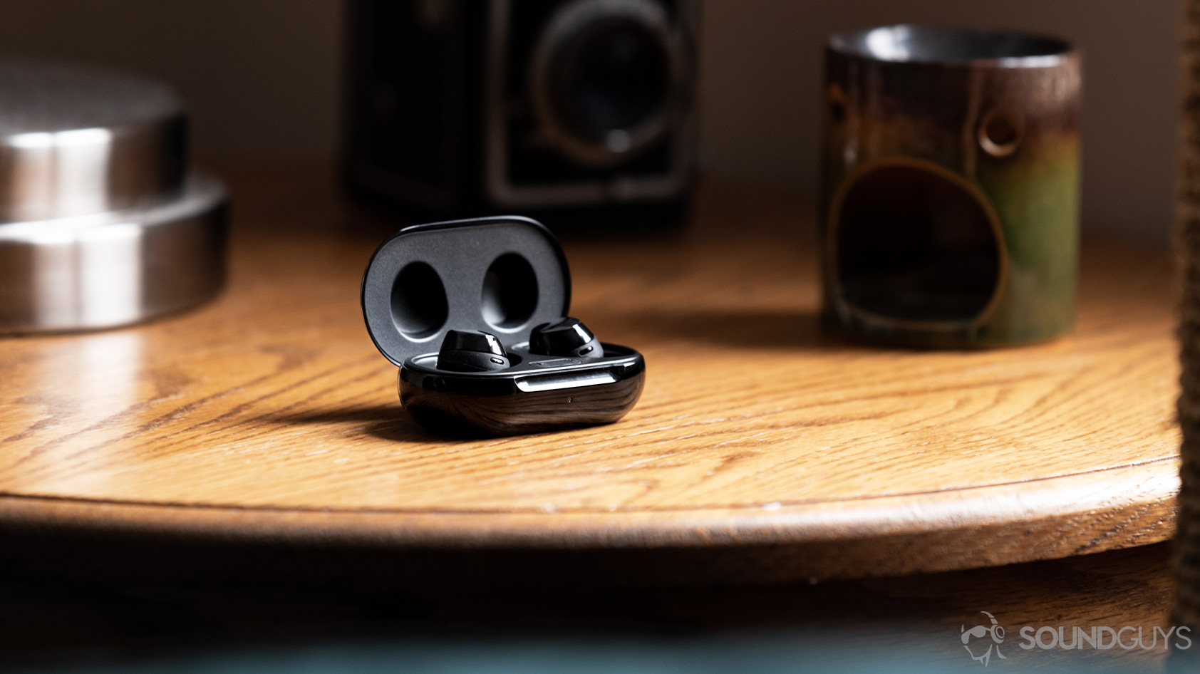 A picture of the Samsung Galaxy Buds Plus true wireless earbuds on a wooden table in front of a vintage camera for the Samsung Galaxy Buds Plus vs Samsung Galaxy Buds Live comparison.