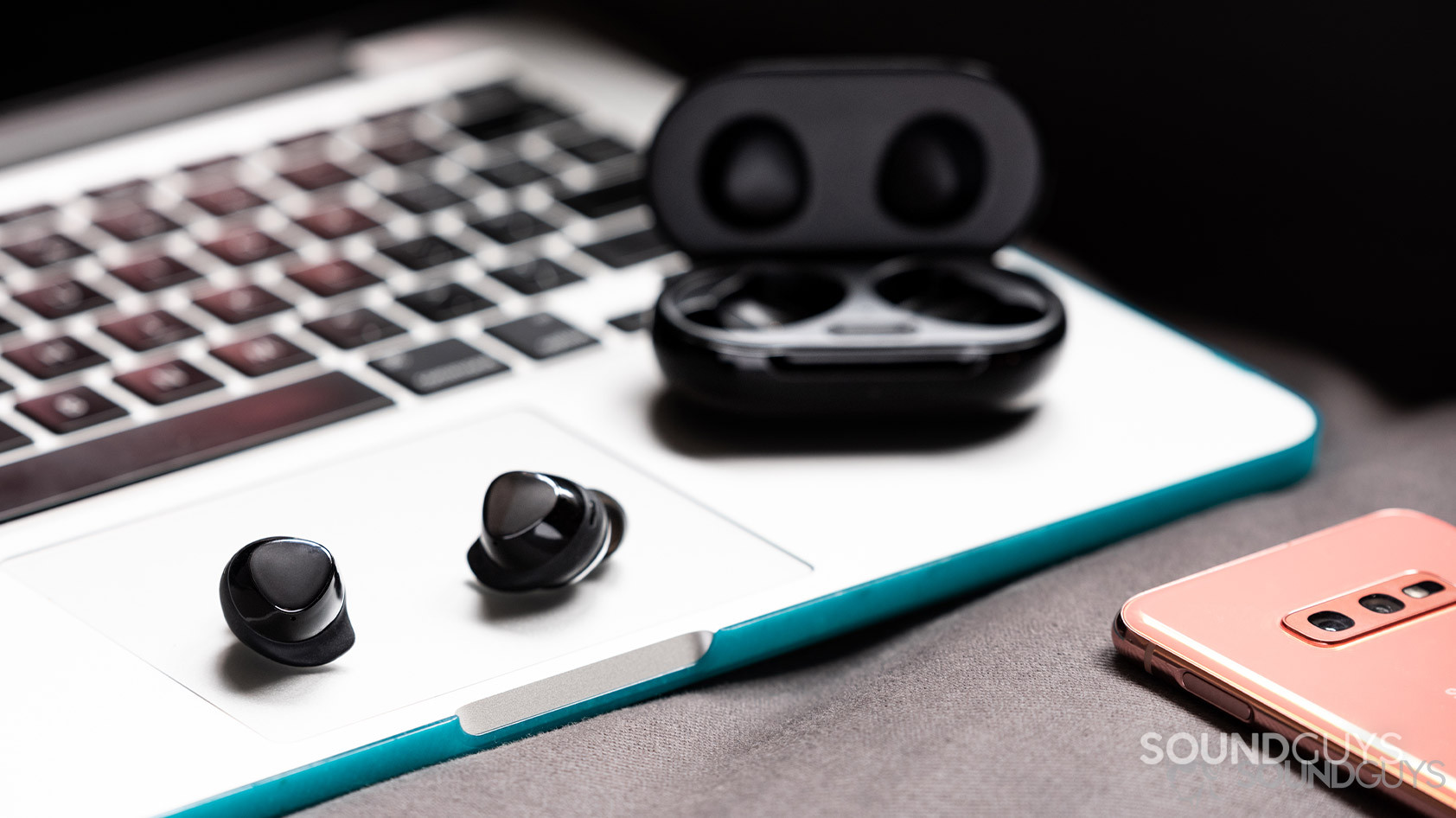 The Samsung Galaxy Buds Plus true wireless earbuds on top of a Macbook Pro with a Samsung Galaxy S10e in the bottom right corner of the image.
