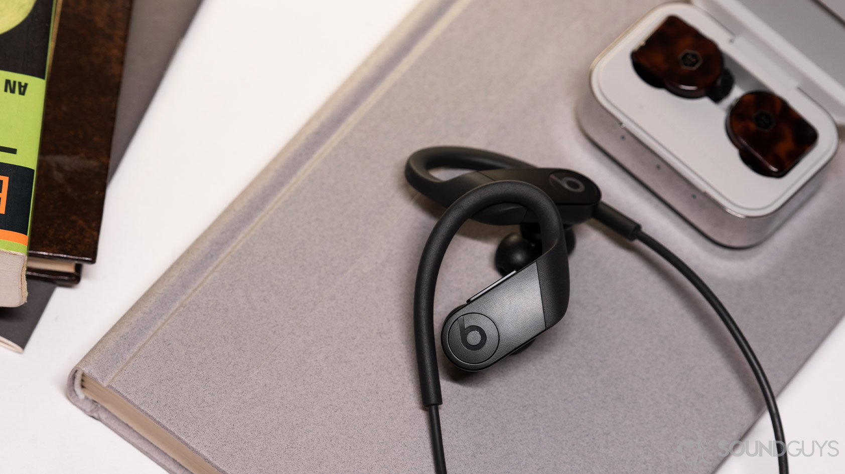 A picture of the Apple Beats Powerbeats on a book and next to the Master &amp; Dynamic MW07 Plus true wireless noise cancellinlg earphones.