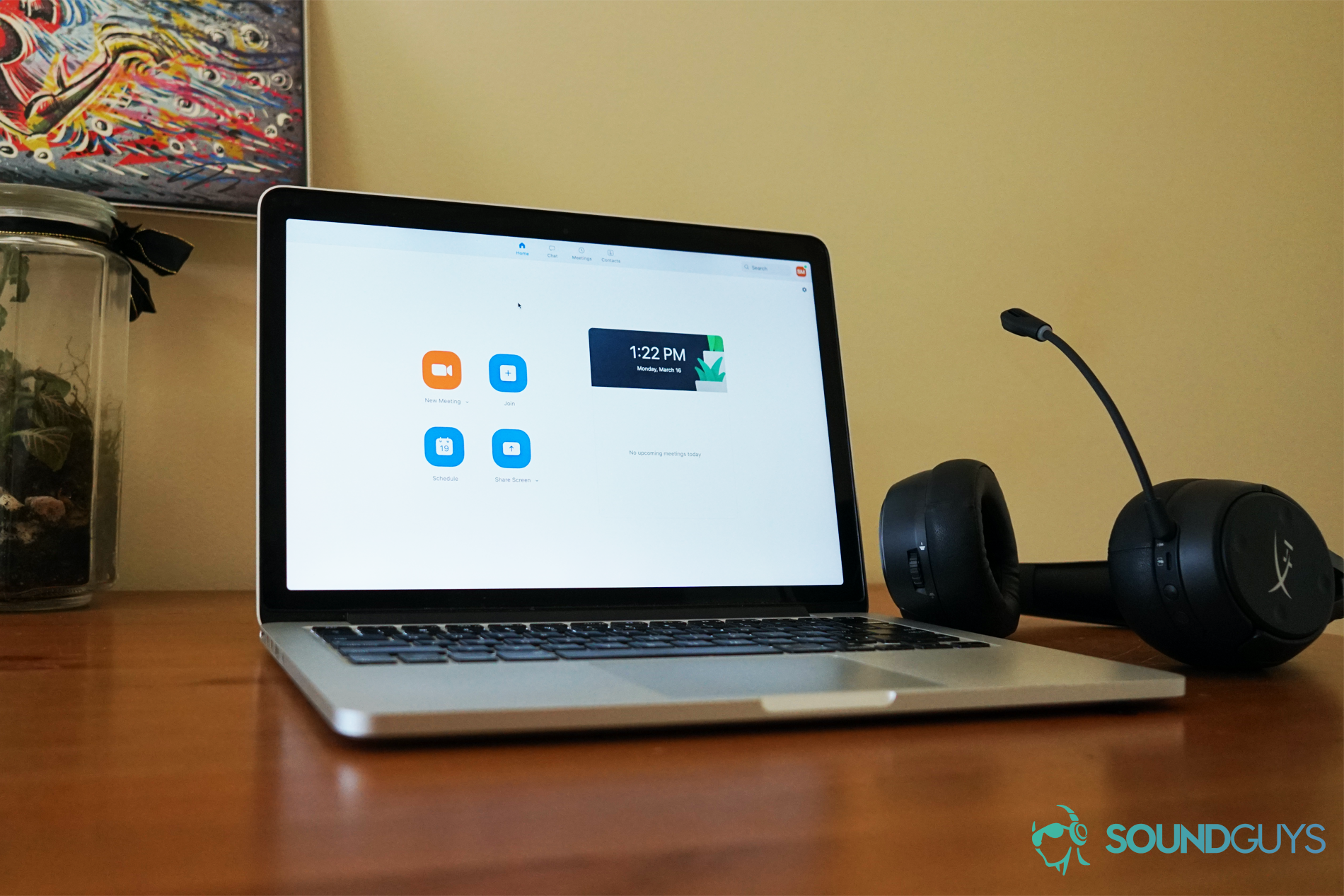 A Macbook Pro running Zoom sits on a wooden table in front of a plant and some art, with a HyperX Cloud Flight S gaming headset next to it.