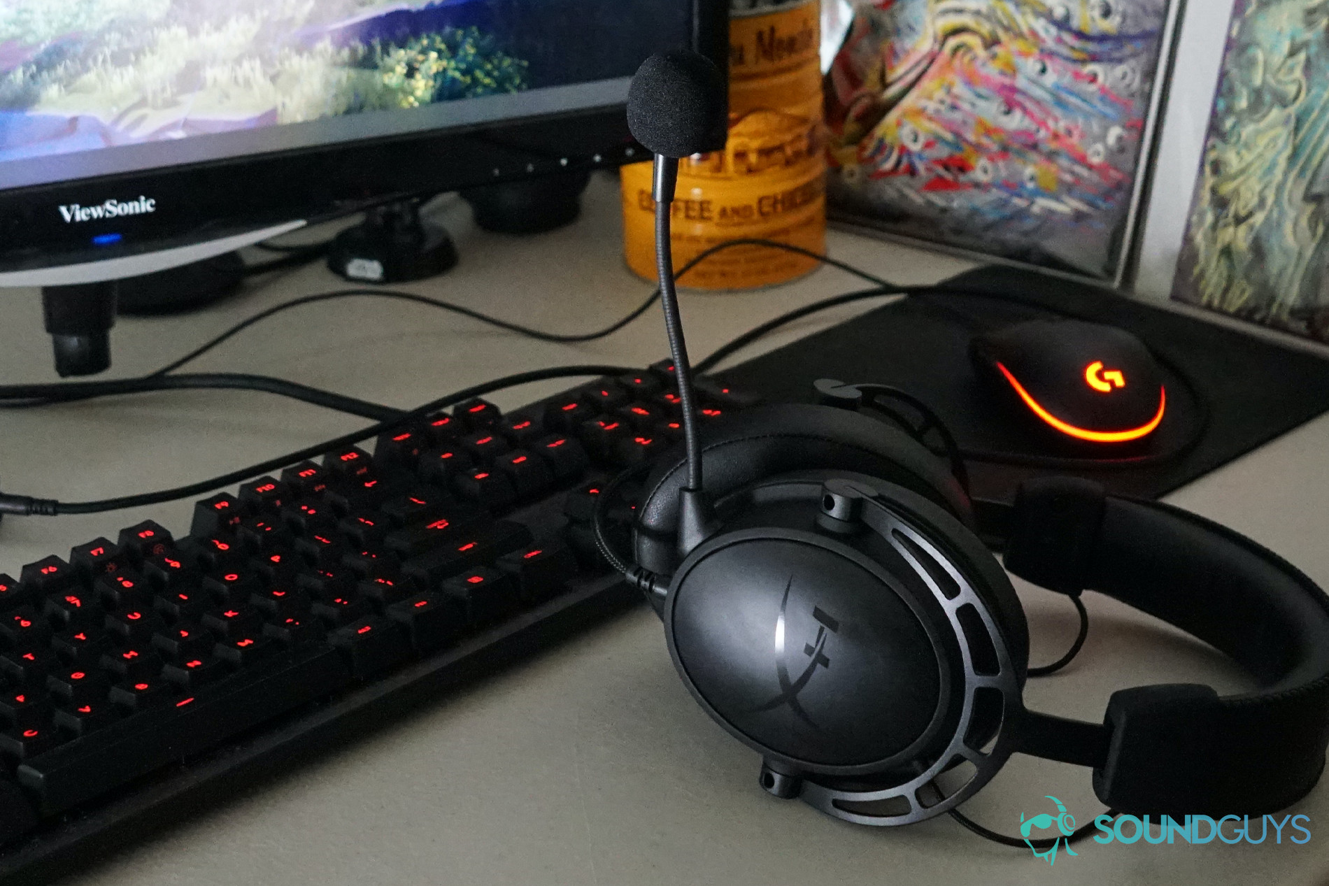 The HyperX Cloud Alpha S sits in front of a gaming PC, in front of a Logitech mouse and gaming keyboard, with tin for Cafe Du Monde coffee and a painting of the Flash in the background.