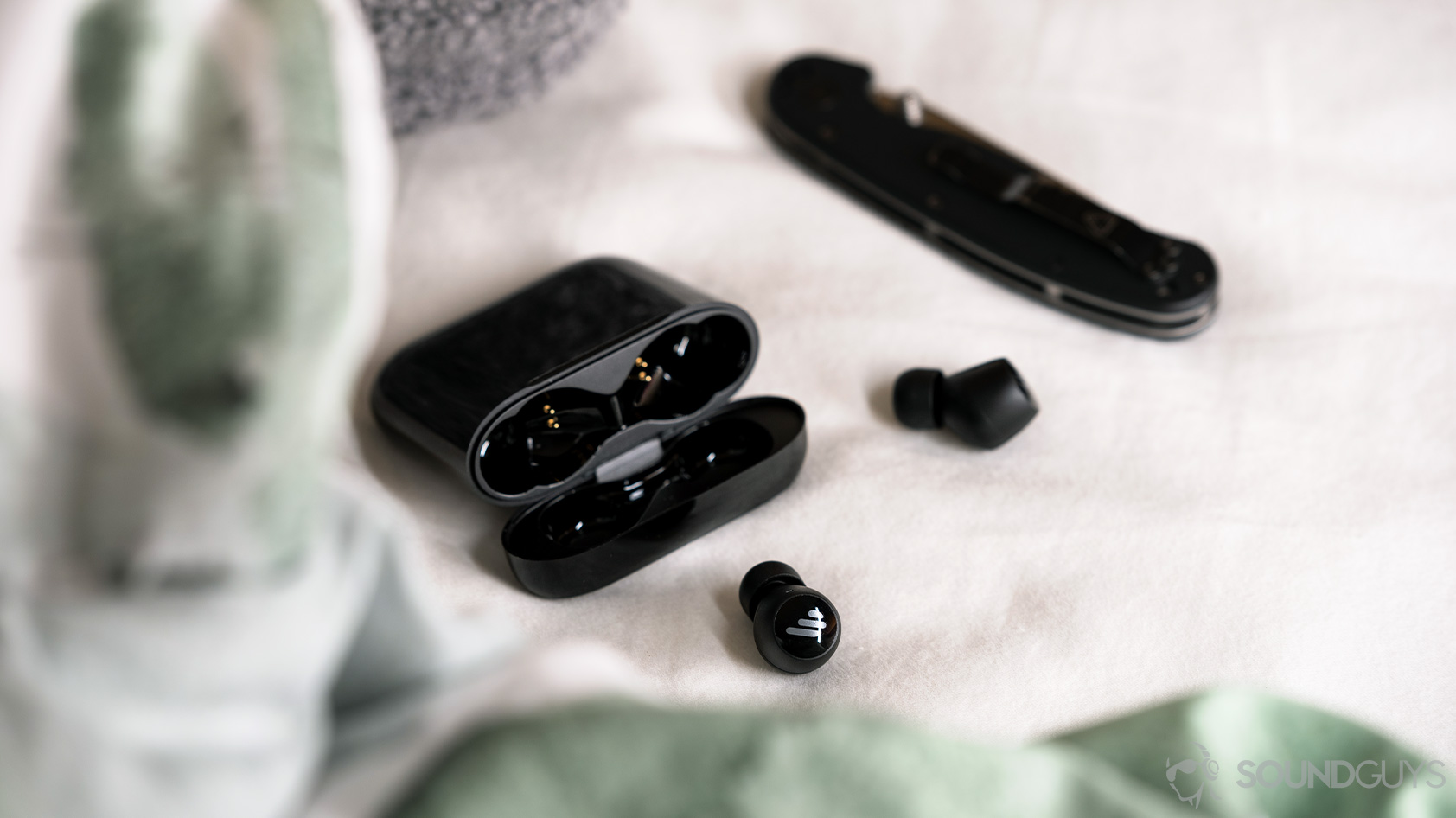 A picture of the Edifier TWS6 true wireless earbuds outside of the open charging case next to a pocket knife on a white bedspread.