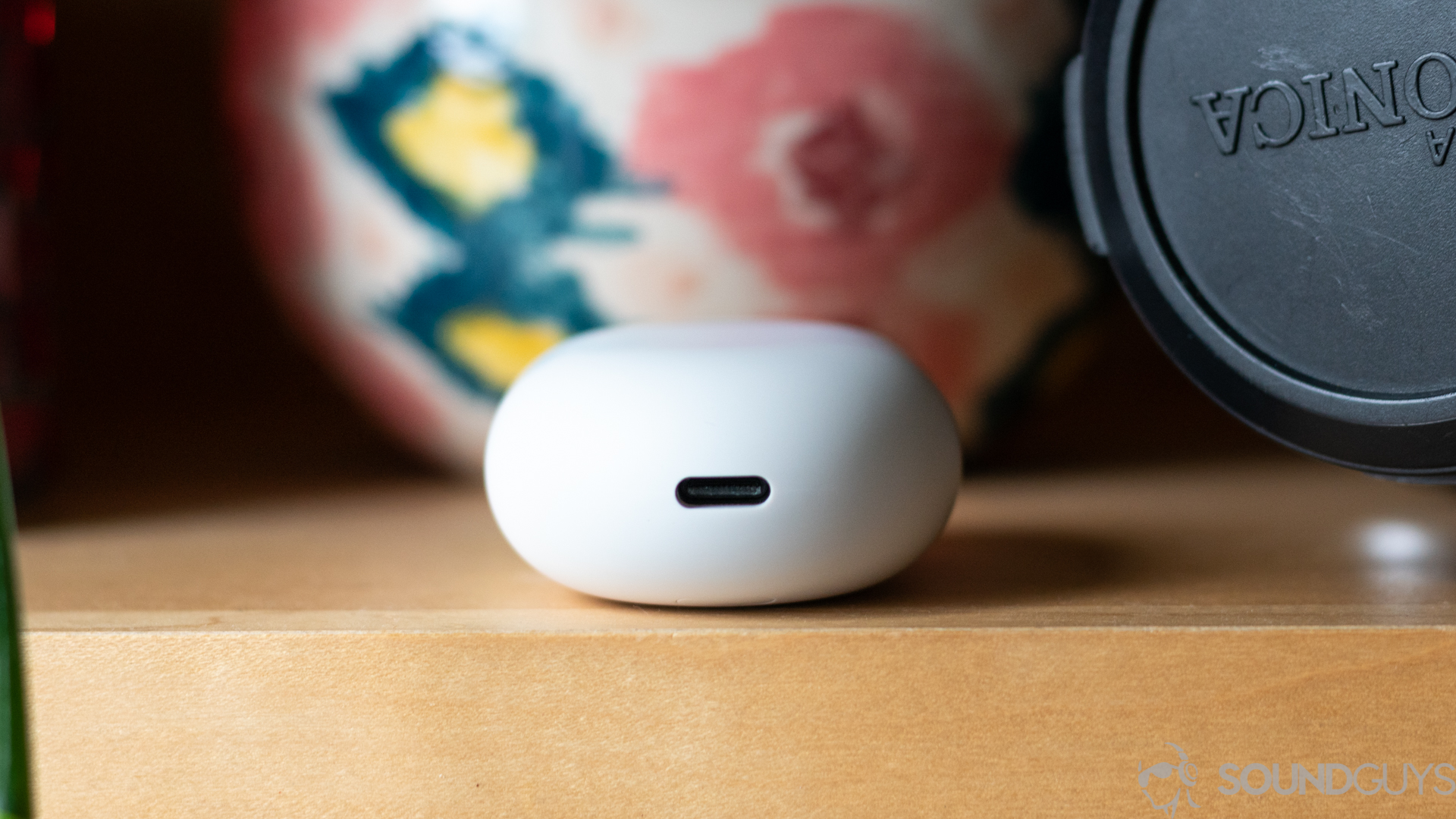 A picture of the Google Pixel Buds (2020) charging case on shelf next to camera and case with close-up of USB-C input on the bottom.