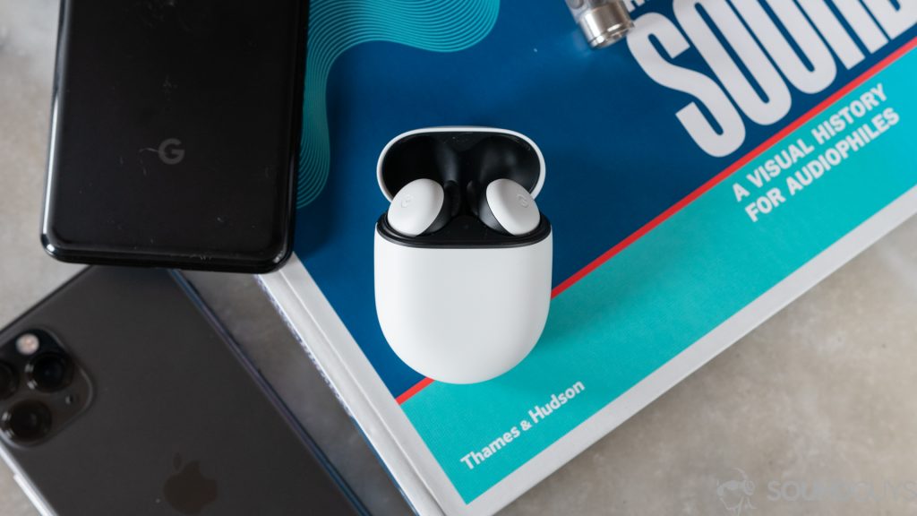 The white new Google Pixel Buds in the open charging case next to both the iPhone 11 Pro and the Pixel 3