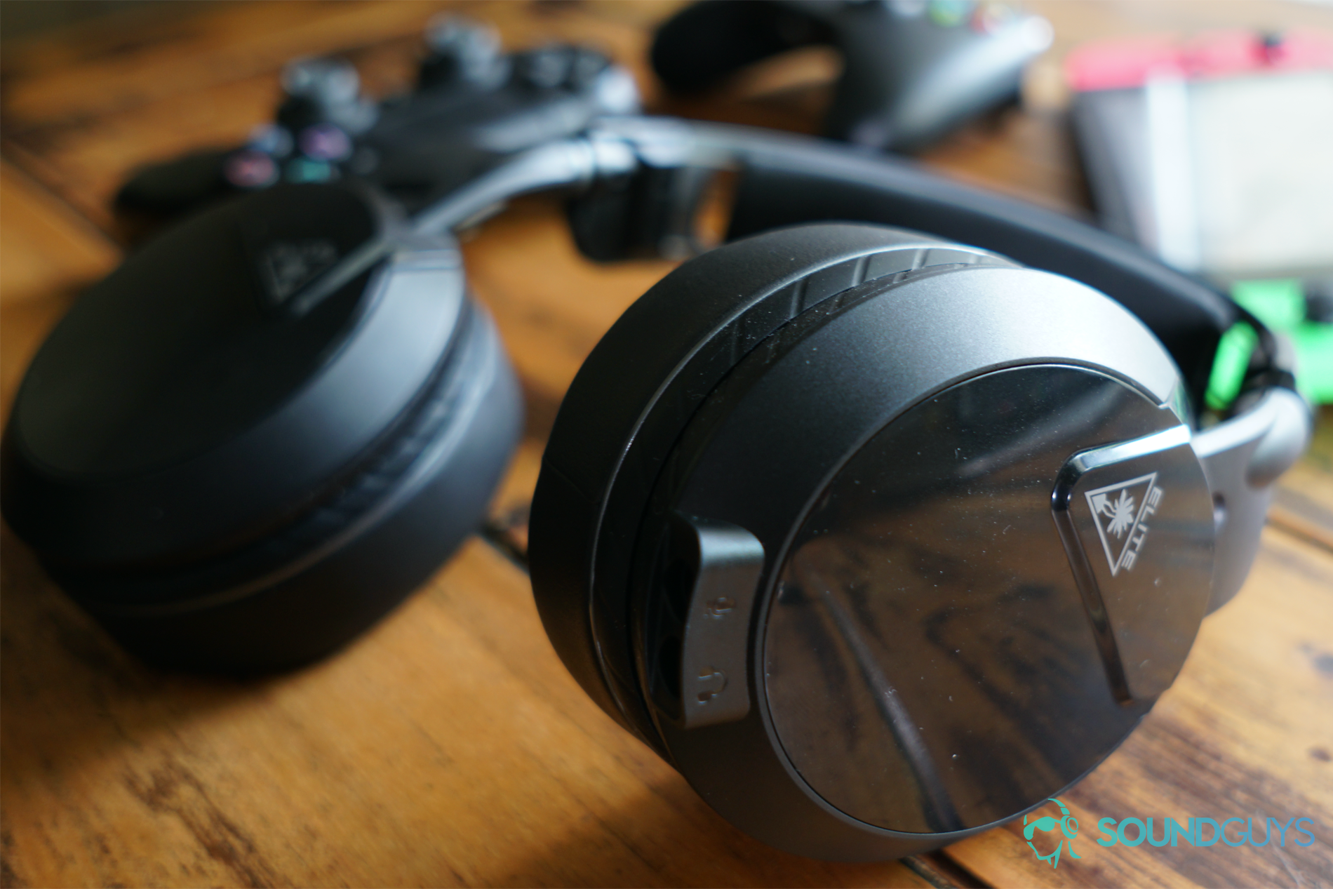The Turtle Beach Elite Atlas gaming headset lays on a wooden table in front of a Nintendo Switch, Xbox One controller, and Playstation 4 Dualshock controller.