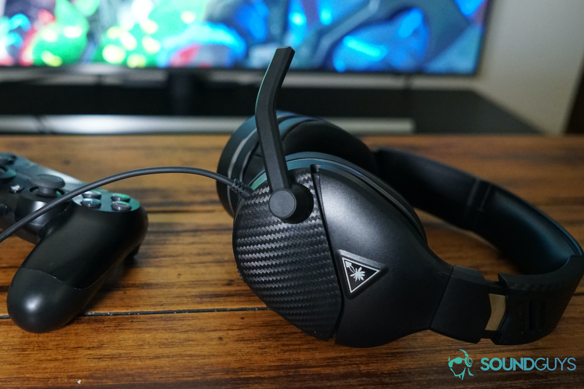 The Turtle Beach Recon 200 gaming headset sits on a table next to a Playstation DualShock 4 controller in front of a television with a Playstation 4 running Dauntless.