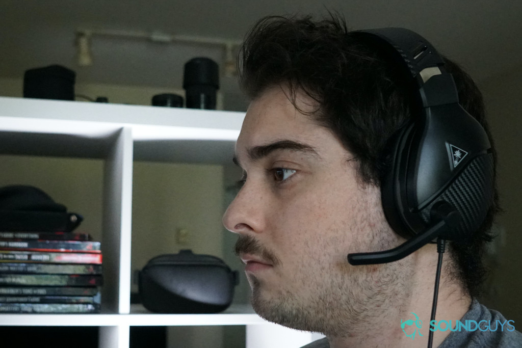 A man wearing the Turtle Beach Recon 200 sits near a shelf with Dungeons and Dragons books and an Oculs Quest on it