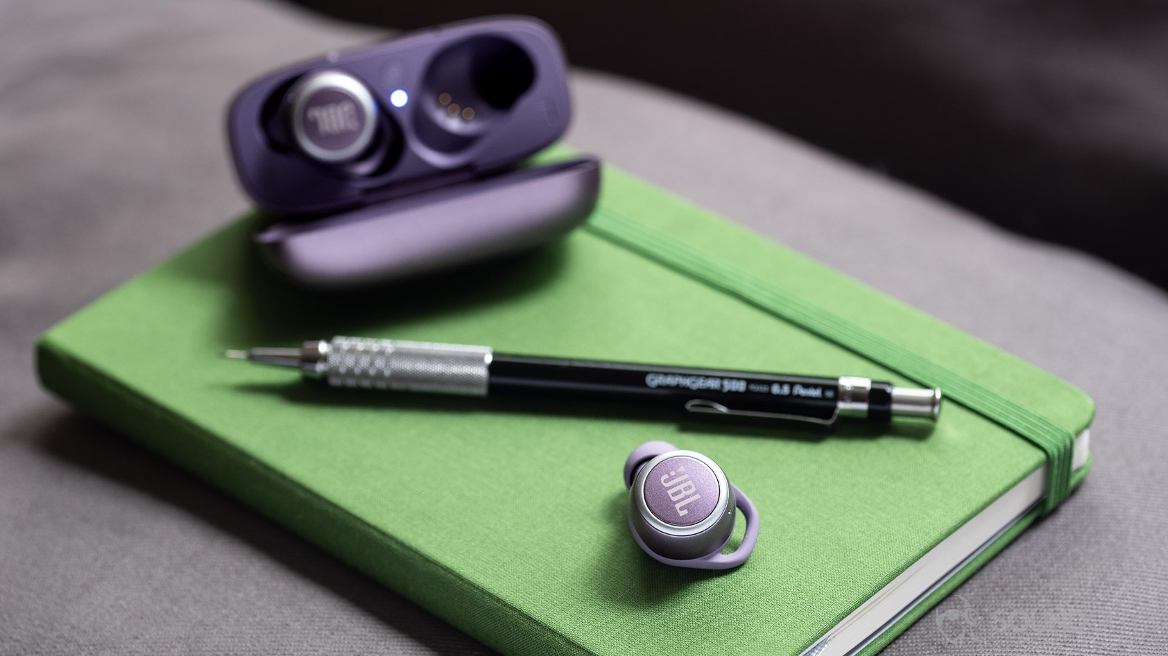 The JBL LIVE 300 TWS true wireless earbuds on a green journal next to a mechanical pencil with the open charging case in the background.