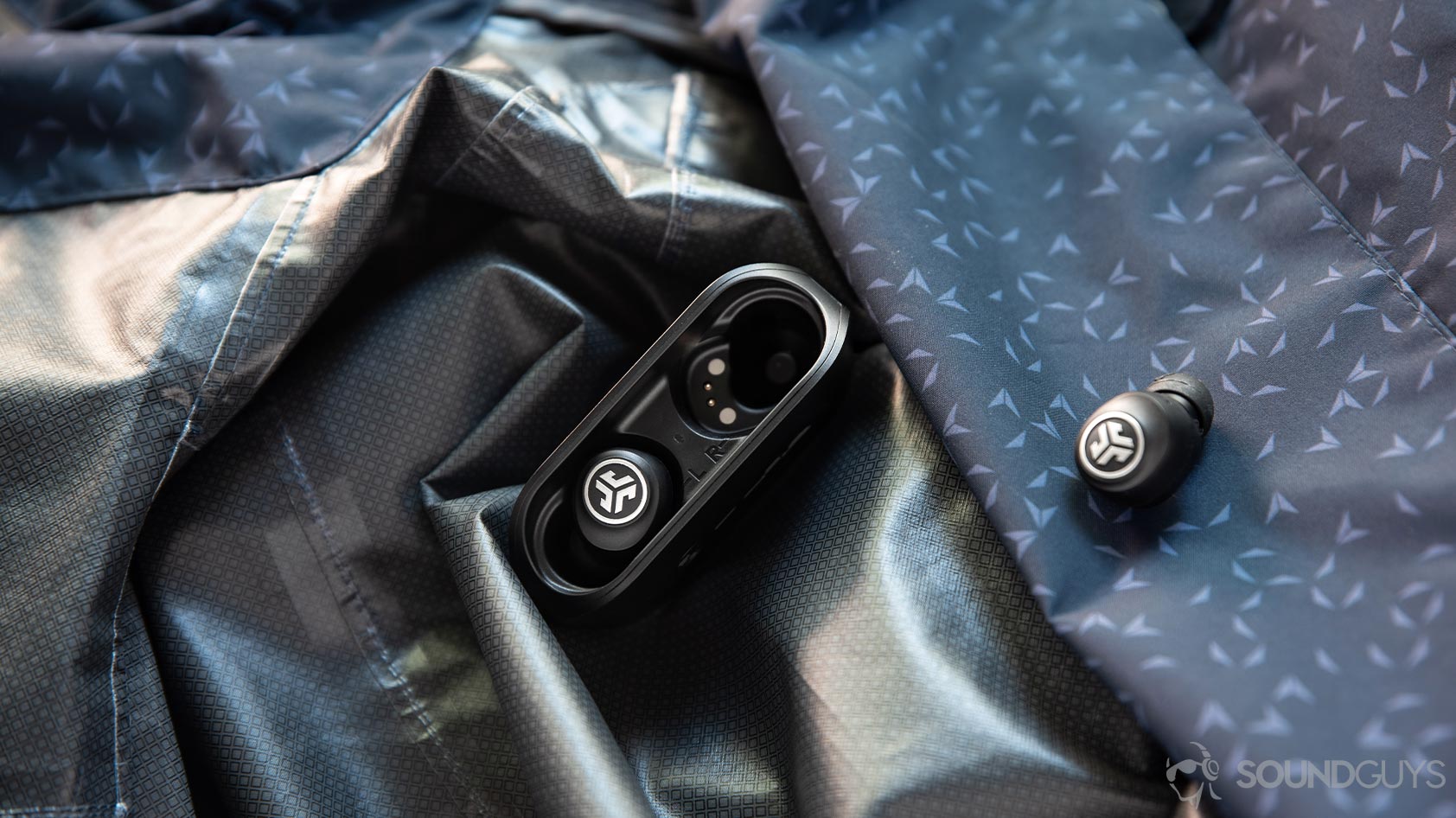 A photo of the JLab GO Air cheap true wireless earbuds on top of a rain jacket with one bud in the case and another outside of it for mono listening.