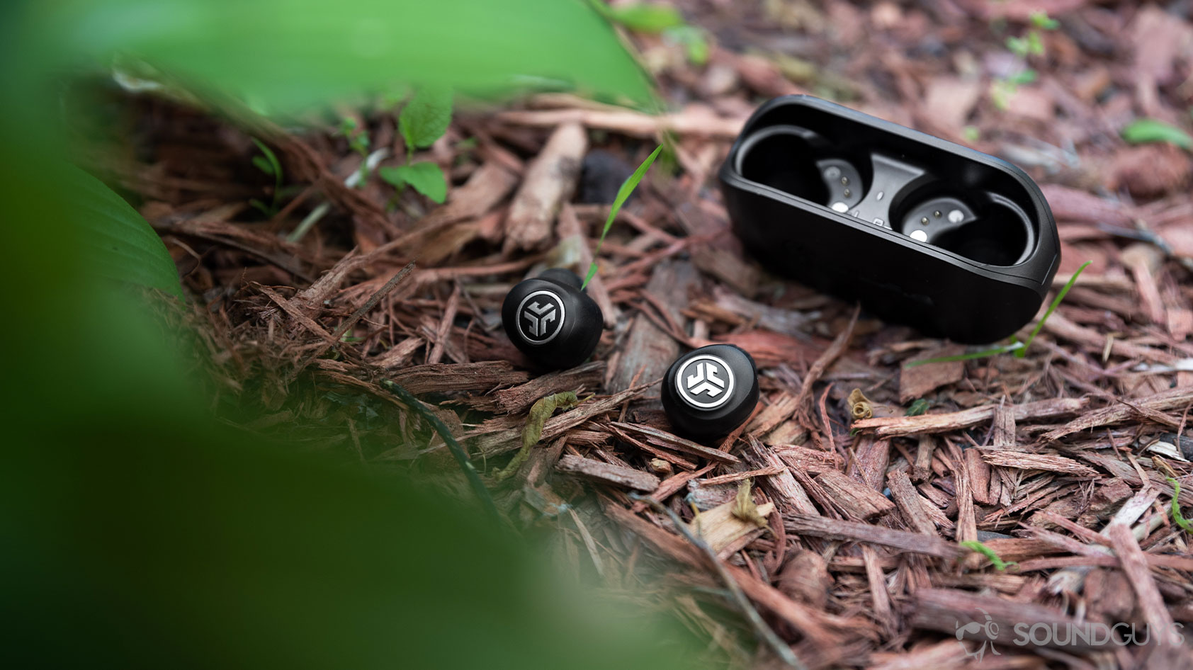 The JLab GO Air cheap true wireless earbuds with the earbuds outside of the case on a mulch pile, almost obscured by leaves in the foreground.