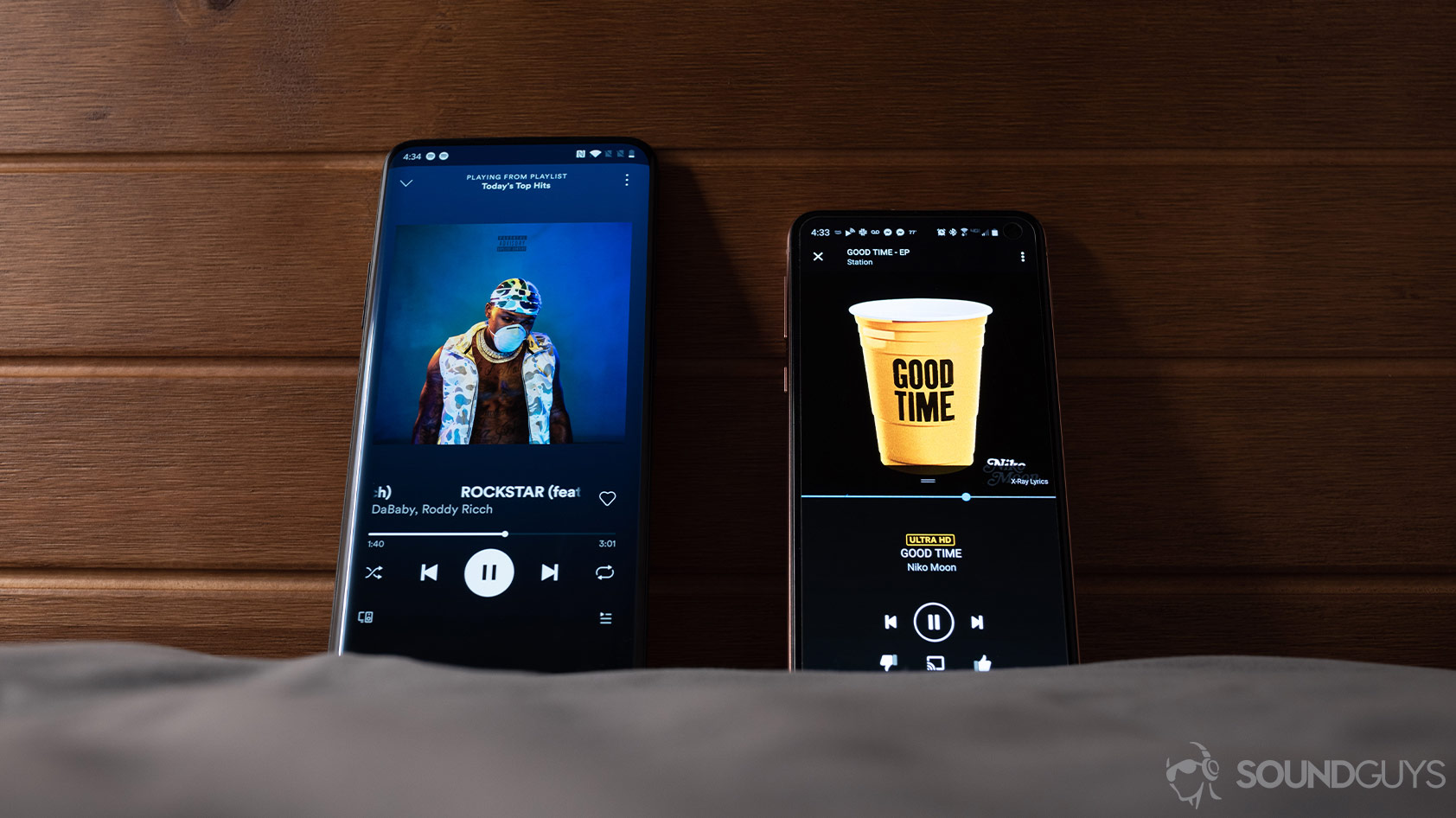 Amazon Music HD vs Spotify Premium music streaming services pulled up on two smartphones against a wood headboard.