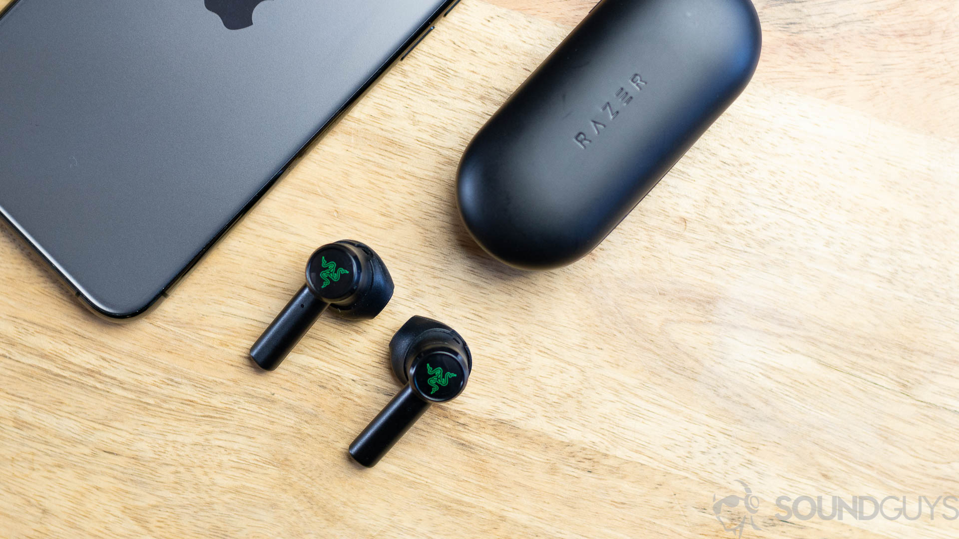 The Razer Hammerhead true wireless earbuds next to iPhone 11 Pro on a wooden surface.