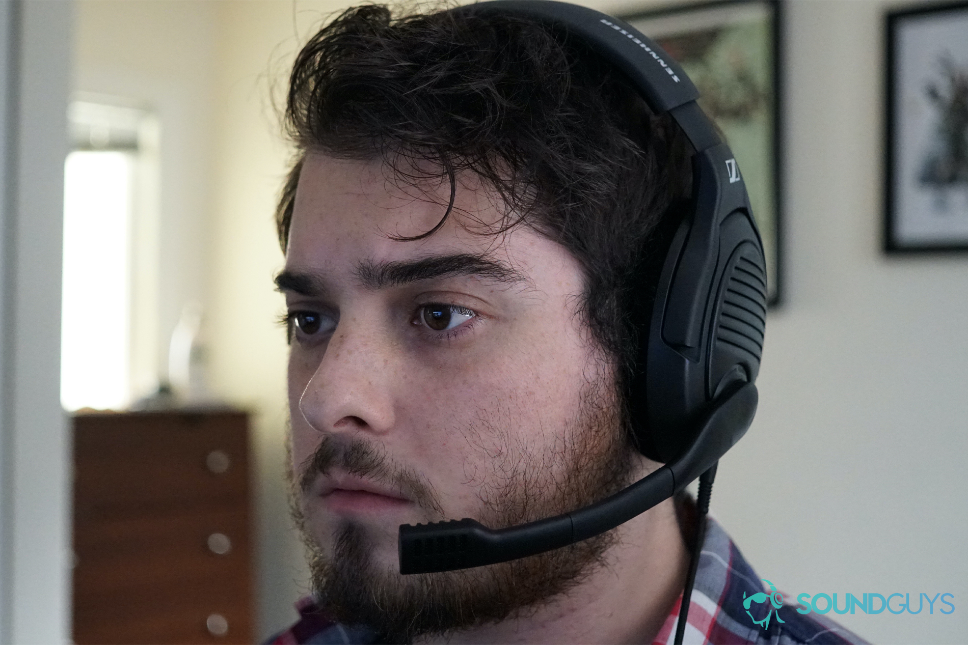 A man wears the Sennheiser PC37X gaming headset while seated at a PC in a room with posters on the wall behind him.