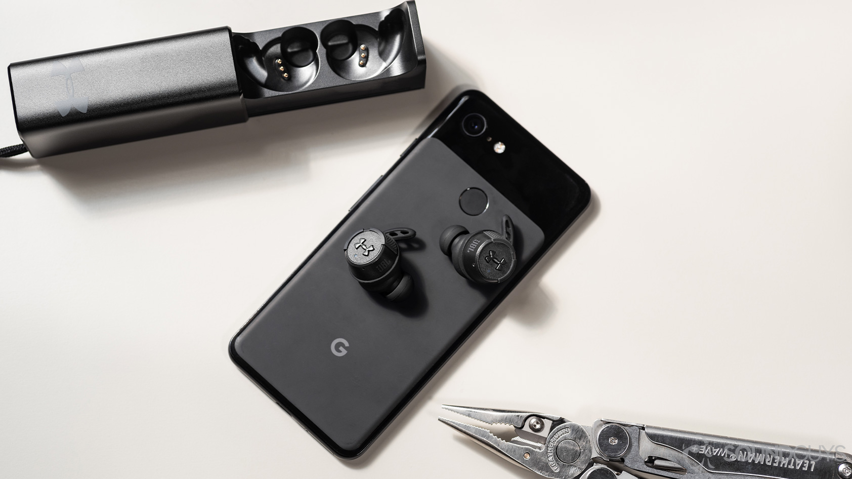 The UA True Wireless FLASH X by JBL on a Google Pixel 3 smartphone, next to the open charging case and a Leatherman Wave+ on a white surface.