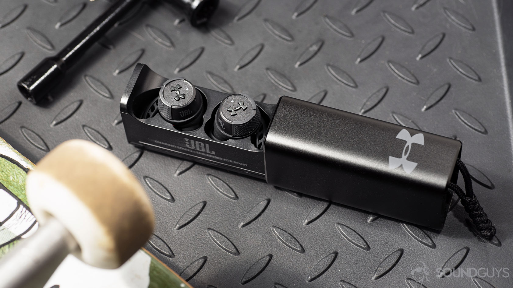 UA True Wireless FLASH X by JBL-workout earbuds case A photo of the UA True Wireless FLASH X by JBL in the open charging case flanked between a skateboard and skateboard tool on a textured surface.