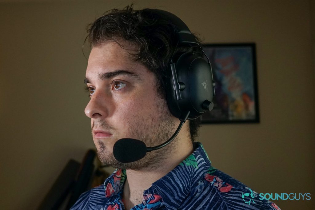 A man wears the Razer BlackShark V2 Pro gaming headset sitting at a PC, with posters for The Adventure Zone and My Brother, My Brother, and Me on the wall behind him.