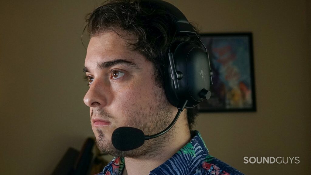 A man wears the Razer BlackShark V2 Pro gaming headset sitting at a PC, with posters for The Adventure Zone and My Brother, My Brother, and Me on the wall behind him.