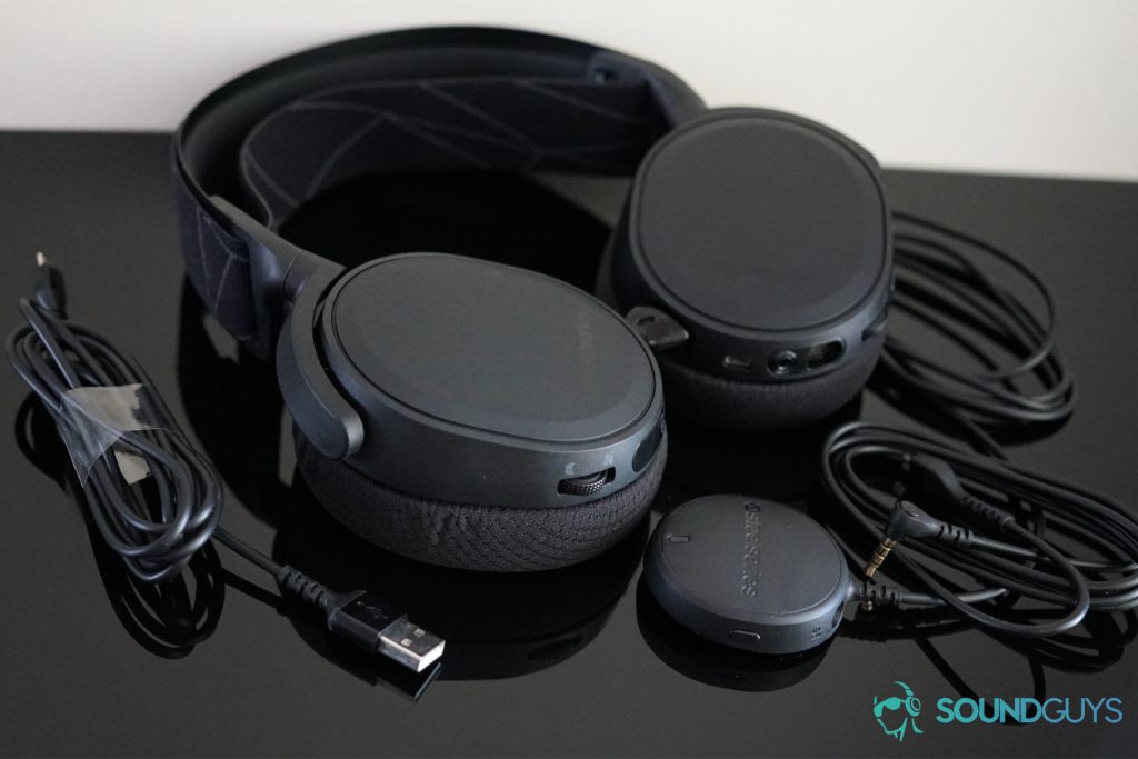 The SteelSeries Arctis 7 Wireless gaming headset lays on a black reflective surface surrounded by all the cords it comes with and its USB wireless dongle.