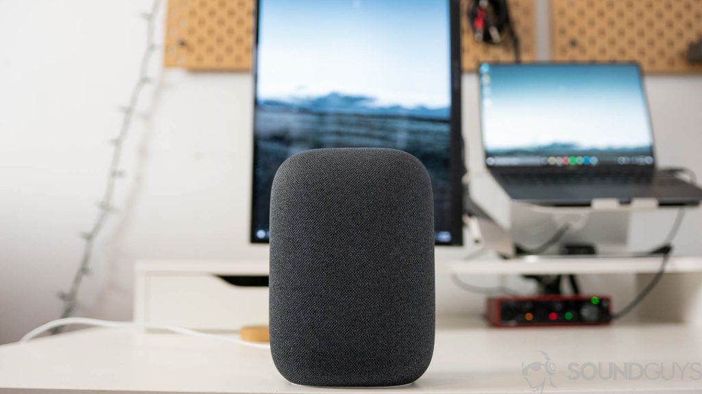The gray Google Nest Audio speaker pictured on a white desk in front of computer screens.