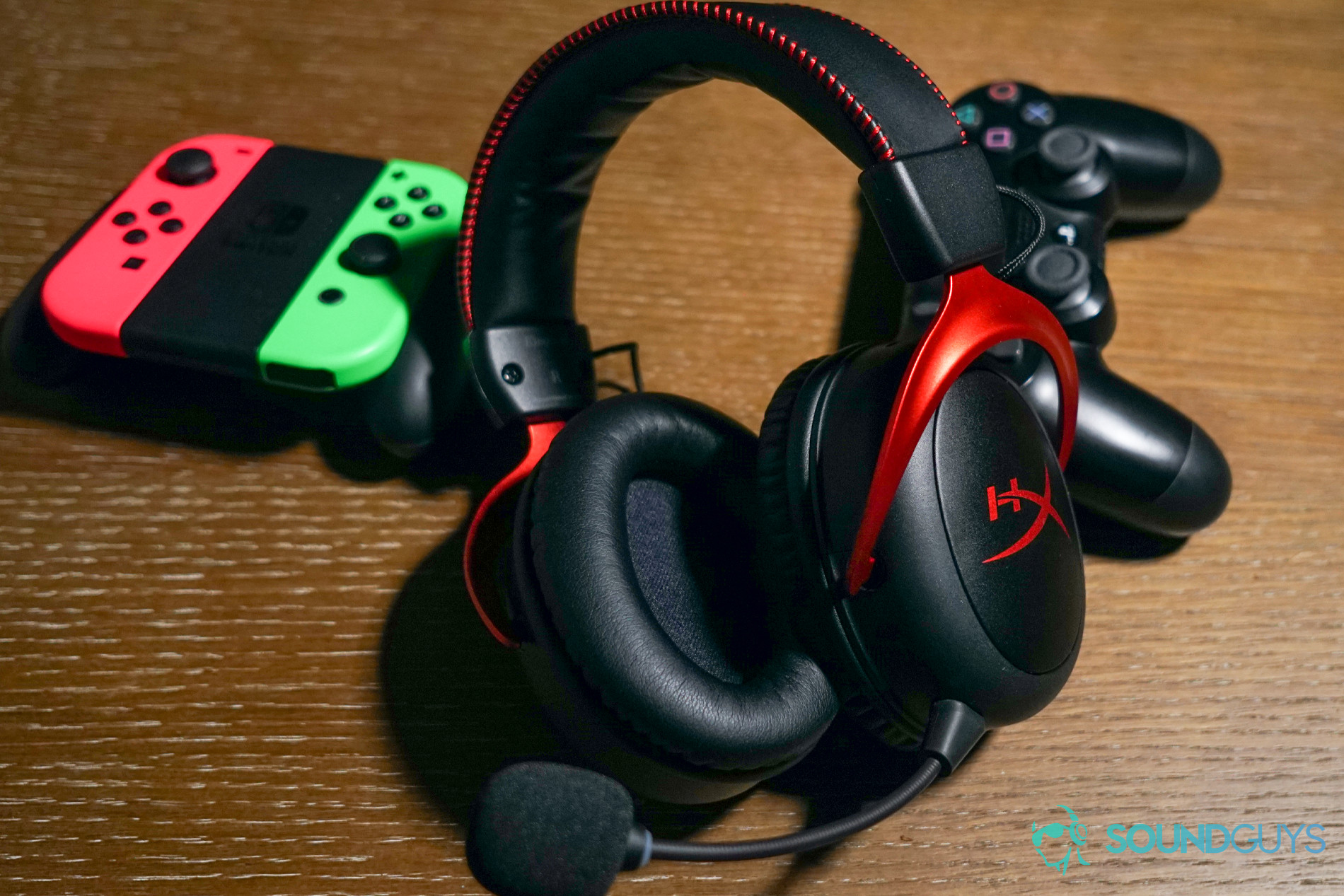 The HyperX Cloud II Wireless lays on a wooden table in front of a PlayStation 4 controller and Nintendo Switch Joy Cons.