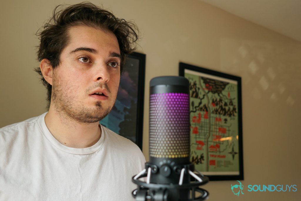 Man positioned behind the HyperX QuadCast S. Wall hangings are visible in the background.