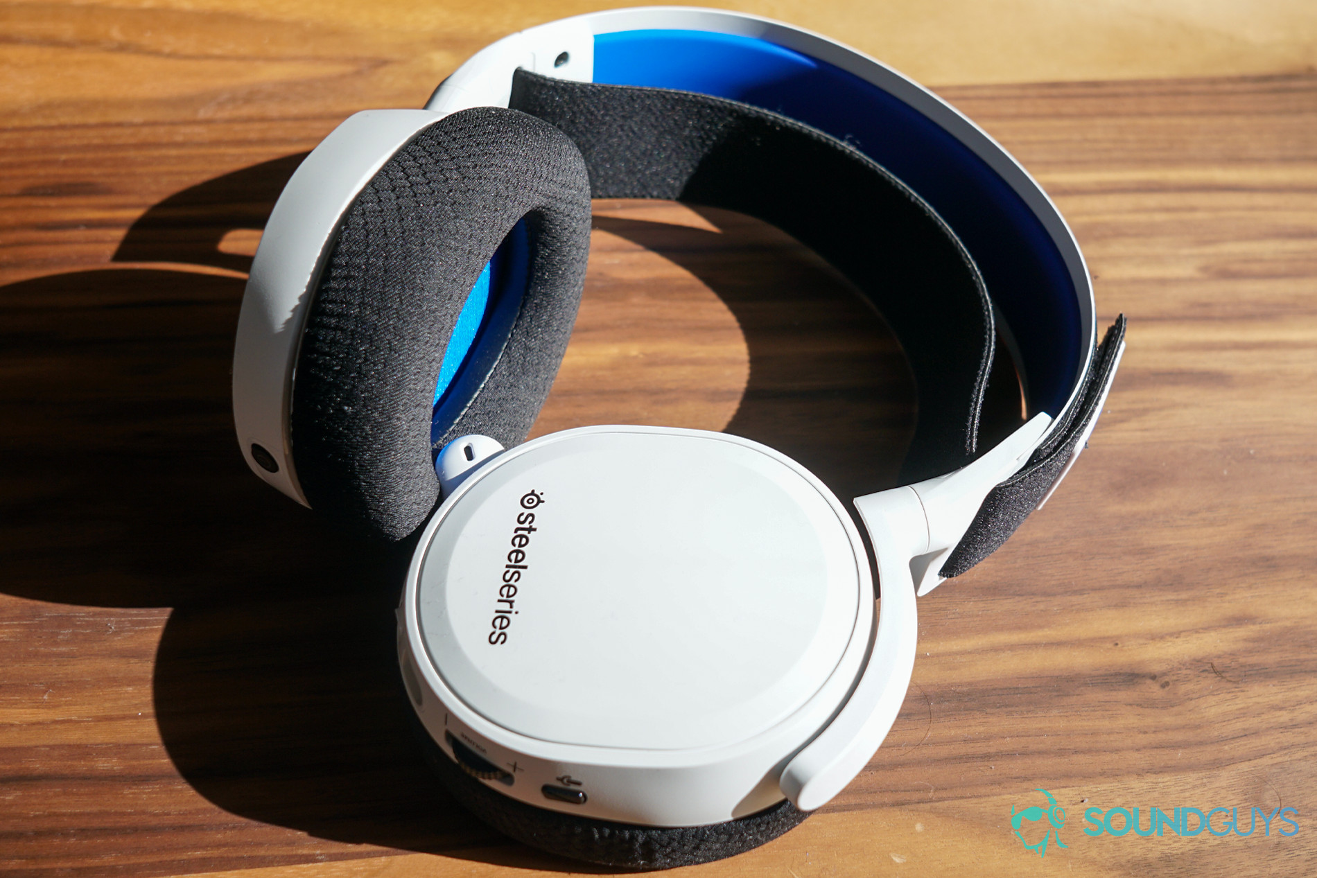 The SteelSeries Arctis 7P gaming headset lays on a wooden table with one headphone rotated flat to the surface.