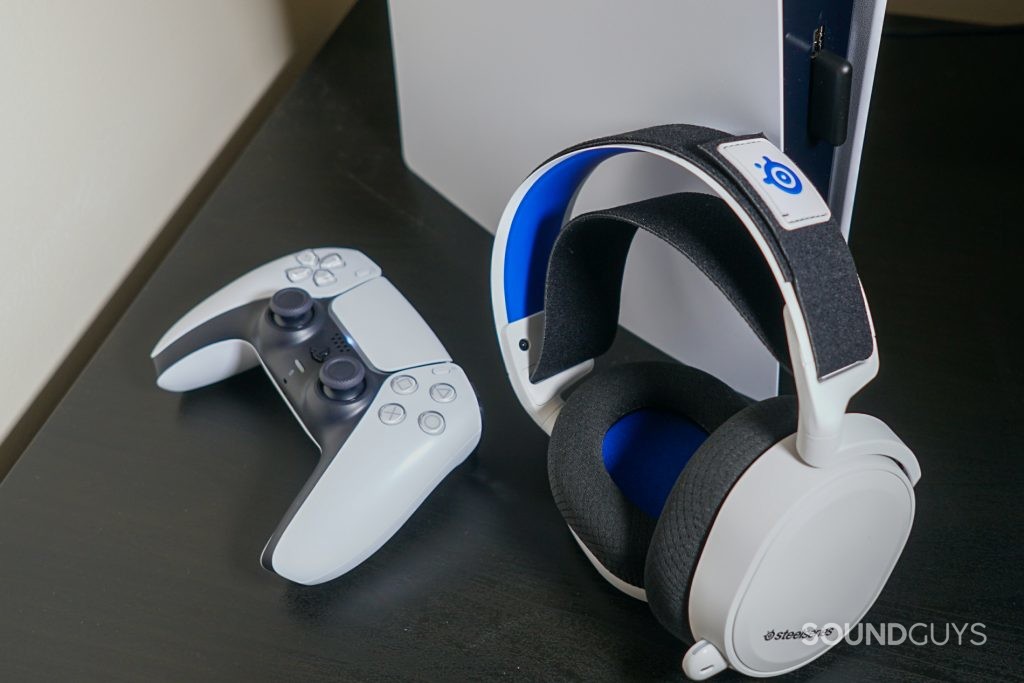 The SteelSeries Arctis 7P leans against a Sony PlayStation 5, next to a PlayStation DualSense controller, with the headset's USB-C dongle plugged into the front of the console.
