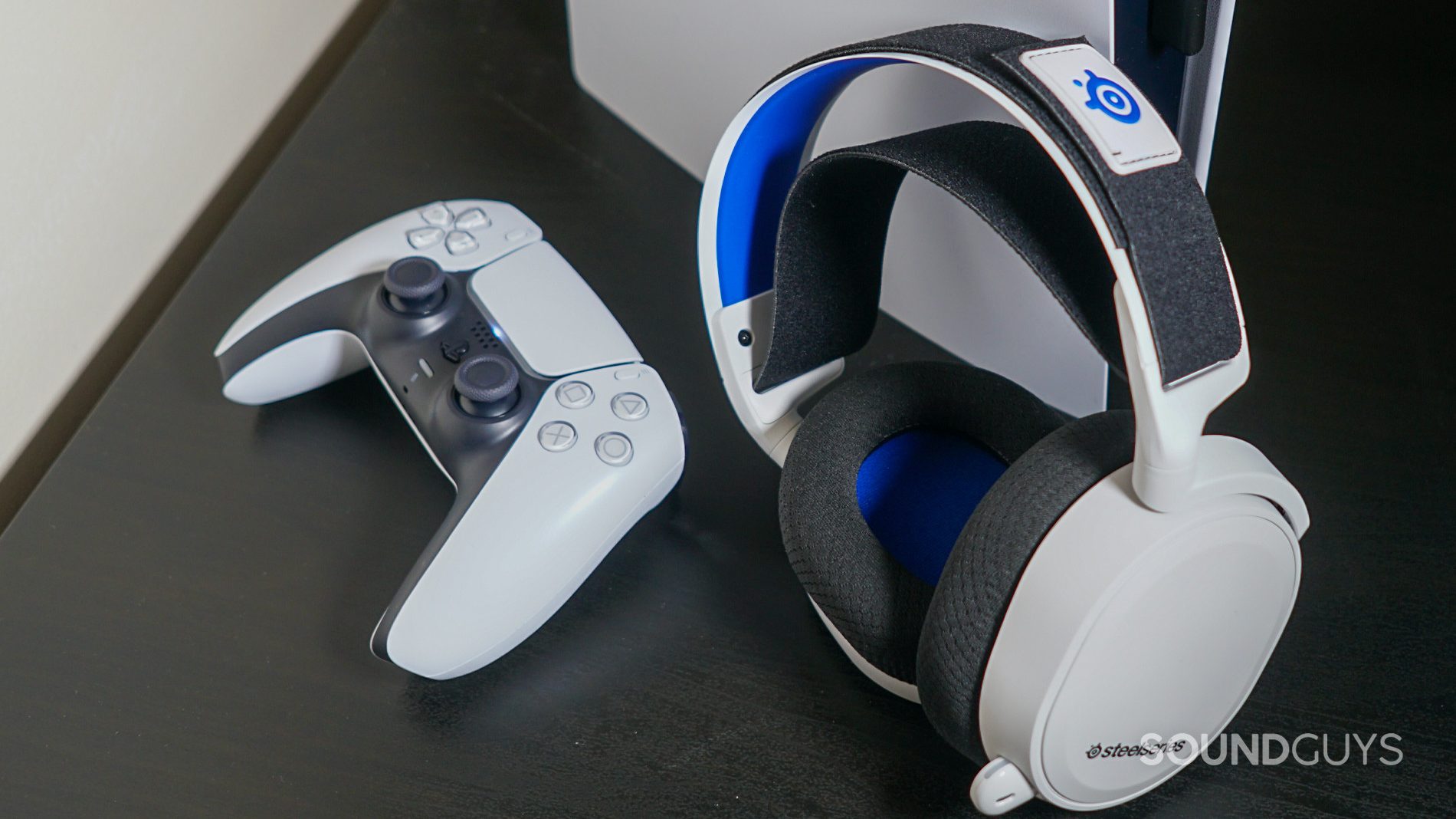 The SteelSeries Arctis 7P leans against a Sony PlayStation 5, next to a PlayStation DualSense controller, with the headset's USB-C dongle plugged into the front of the console.