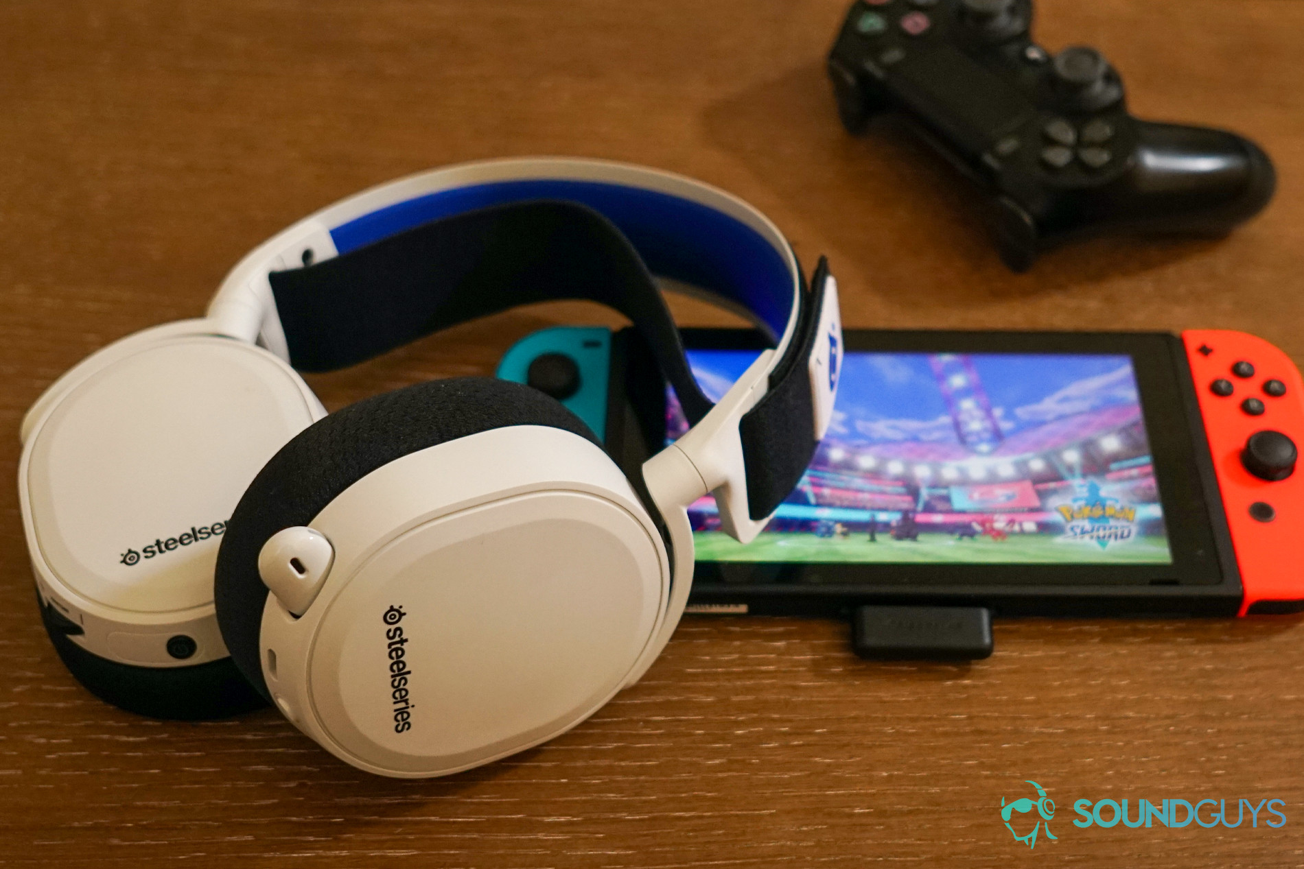 The SteelSeries Arctis 7P lays on top of a Nintendo Switch running Pokemon Sword, which its wireless USB-C dongle is plugged into, with a PlayStation DualShock 4 controller in the background.