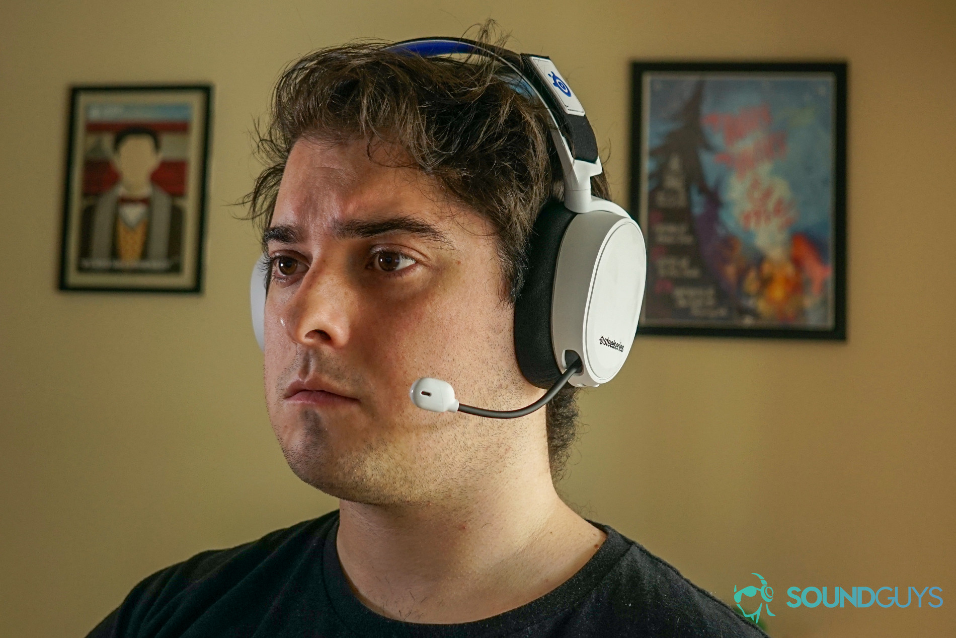 A man wears The SteelSeries Arctis 7P gaming headset in front of posters for My Brother, My Brother, and Me, and a Canadian Heritage Minute 