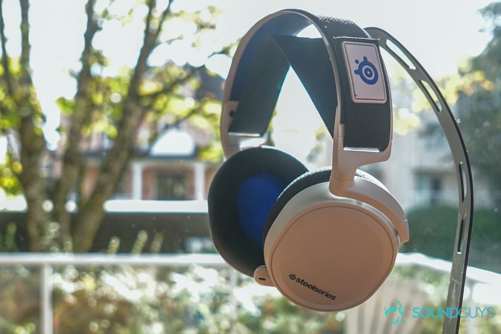The SteelSeries Arctis 7P hangs from a headphone stand in front of a window