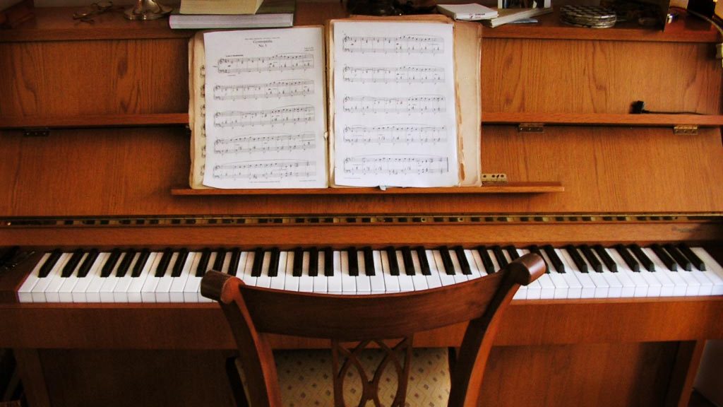 A light brown piano with sheet music on its stand. A chair is placed in front of the keys.